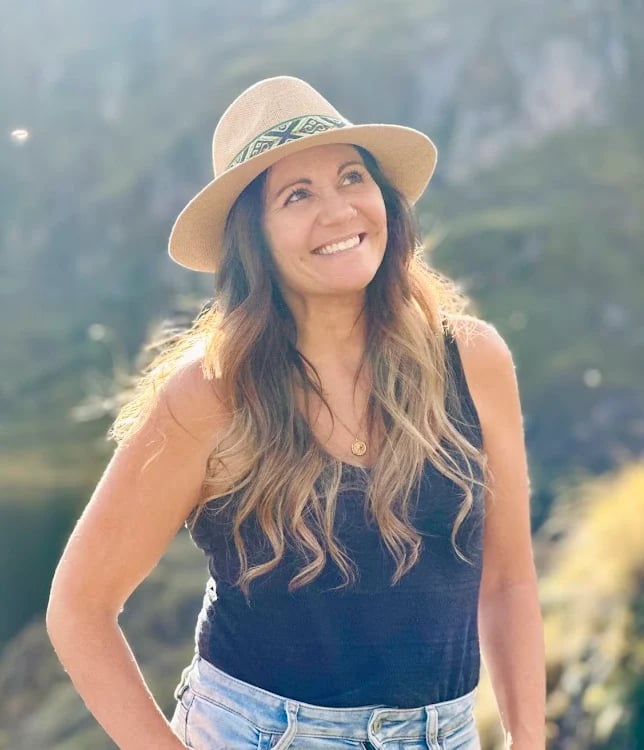 Smiling woman with long wavy hair wearing a tan cowboy hat and denim outfit in a sunny mountain landscape