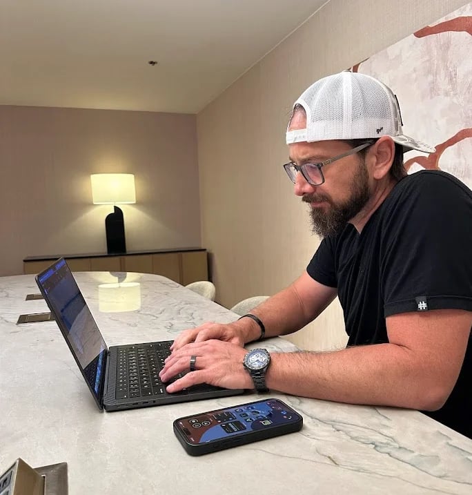 Man with beard and white cap working on laptop at marble desk in modern bedroom with smartphone nearby